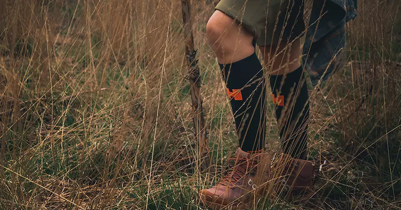 Een persoon die in hoog gras staat, gekleed in zwarte kniekousen met oranje details, bruine laarzen en een korte broek. Alleen de onderste helft van het lichaam is zichtbaar.
